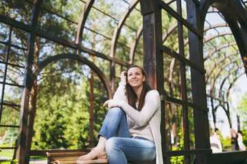 Portrait of beautiful caucasian young woman with charming smile wearing light casual clothes. Smiling female sitting under archway in city park in street outdoors on spring nature. Lifestyle concept.