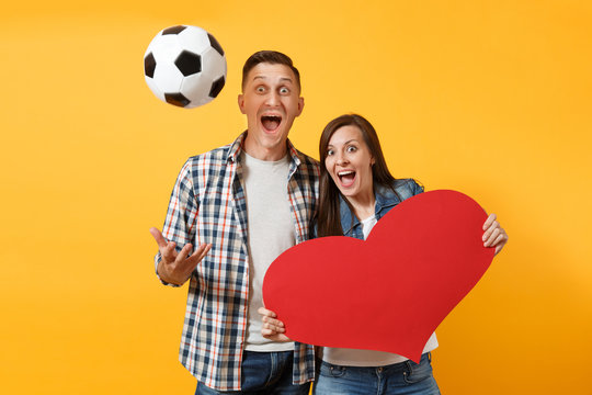 Young happy cheerful couple supporter, woman man, football fans cheer up support team, holding red heart love, soccer ball isolated on yellow background. Sport family leisure people lifestyle concept
