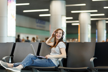 Young laughing traveler tourist woman with headphones listening music working on laptop, wait in lobby hall at international airport. Passenger traveling abroad on weekend getaway. Air flight concept.