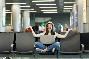 Fototapeta na wymiar Young joyful traveler tourist woman with laptop sitting with crossed legs, meditate, spread hands, waiting in lobby hall at airport. Passenger traveling abroad on weekend getaway. Air flight concept.