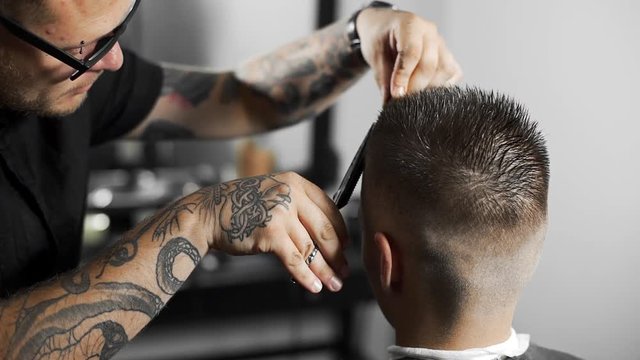 Tattoed barber makes haircut for customer at the barber shop by using scissors and comb , man's haircut and shaving at the hairdresser, barber shop