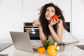 Photo of young curly woman 20s holding sweet paper and looking upward, while using silver laptop for cooking dinner in kitchen interior