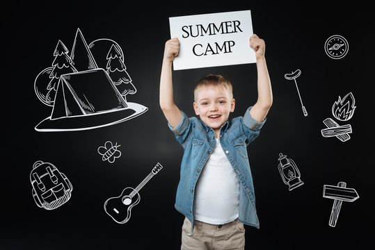 Amazing summer camp. Enthusiastic active boy feeling good and putting a sign about summer camp up while relaxing and enjoying the beauty of nature