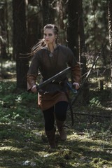 Viking woman with sword and hammer wearing traditional warrior clothes in a deep mysterious forest.