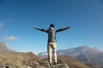 an applause of a young woman backpacker on top of a sprawling upward mountain peak. Freedom and victory against the background of mountains and blue sky