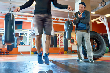 Crop man jumping rope in boxing gym under control of elderly man with timer standing near.