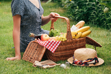 cropped image of woman sitting on green grass at picnic and touching basket with food