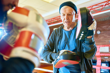 Crop man in gloves practicing box punches with senior trainer holding focus mitts ad giving...