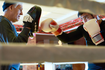 Side view of serious man boxing with senior coach holding special training pads on ring. 