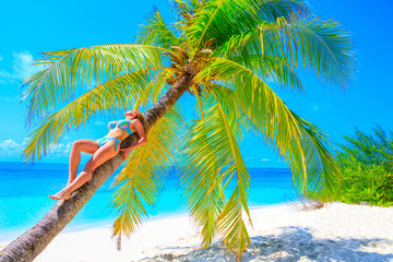 Gorgeous blond woman in bikini basks on the palm tree on the sandy beach