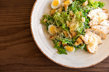 Fresh summer salad Ceasar with eggs, lettuce, chicken and parmesan cheese on white plate on wooden background; healthy concept; top flatlay view