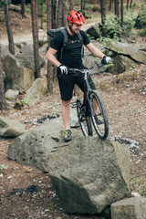 Fototapeta premium smiling male extreme cyclist in protective helmet standing with mountain bicycle on stone in forest