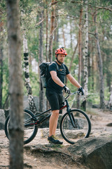 young male extreme cyclist in protective helmet standing with mountain bicycle in forest