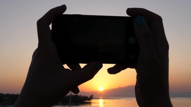 Female hands hold mobile phone silhouette while take picture of picturesque sunset in Greece