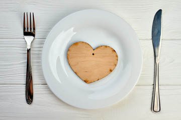 White plate with wooden heart decoration. Plywood heart on plate, silver fork and knife on white wooden table. Table setting for Valentines holiday.