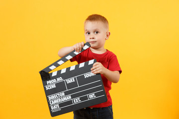 Little cute kid baby boy 3-4 years old in red t-shirt holding in hand classik black film making clapperboard looking camera isolated on yellow background. Kids childhood lifestyle concept. Copy space.