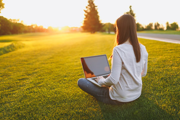 Young female back view. Woman working on laptop pc computer with blank black empty screen to copy space in city park on green grass sunshine lawn outdoors. Mobile Office. Freelance business concept.