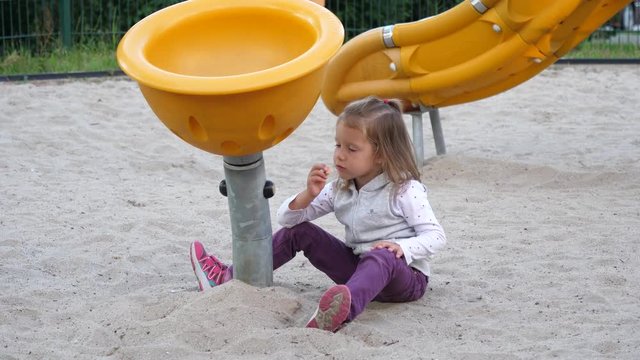 Germany little kid girl sit on a sand in sandbox of playground eating chewing cookies