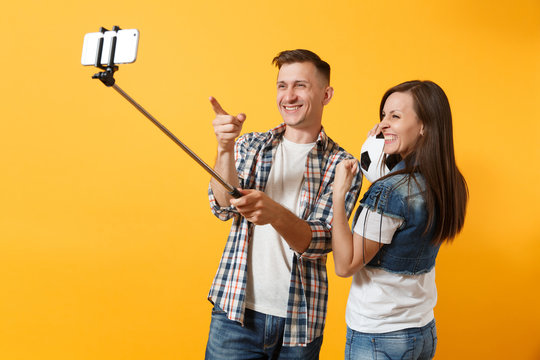 Young couple, woman man, football fans doing selfie on mobile phone with monopod selfish stick, cheer up support team, soccer ball isolated on yellow background. Sport family leisure lifestyle concept