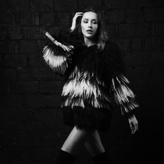 Full body portrait of gorgeous brunette woman wearing fashionable faux fur coat with long black and white hair and over knee boots. Seductive female model posing against gray brick wall on background.