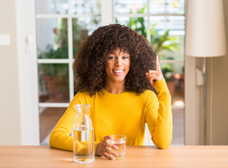 African american woman drinking a glass of water at home surprised with an idea or question pointing finger with happy face, number one