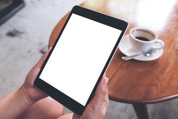 Mockup image of a woman sitting and holding black tablet pc with white blank screen with coffee cup on table