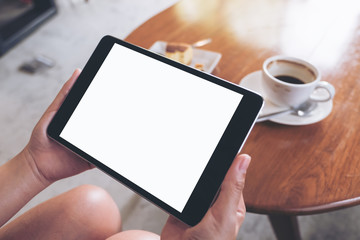 Mockup image of a woman sitting and holding black tablet pc with white blank screen with coffee cup on table