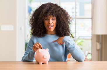 African american woman saves money in piggy bank with surprise face pointing finger to himself