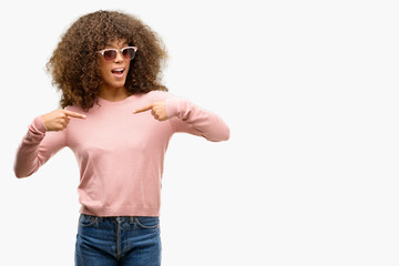African american woman wearing pink sunglasses looking confident with smile on face, pointing...