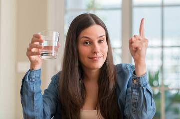 Young woman drinking glass of water at home surprised with an idea or question pointing finger with...