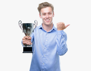 Young handsome blond man holding a trophy pointing with hand and finger up with happy face smiling