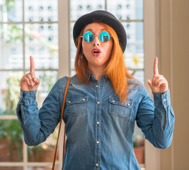 Stylish redhead woman wearing bowler hat and sunglasses amazed and surprised looking up and...