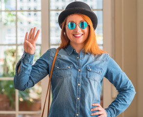 Stylish redhead woman wearing bowler hat and sunglasses showing and pointing up with fingers number...