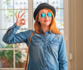 Stylish redhead woman wearing bowler hat and sunglasses smiling positive doing ok sign with hand...
