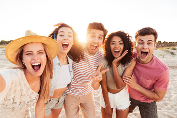 Photo of joyful young multiracial people in summer clothes screaming, while taking selfie during walk near sea at sunset