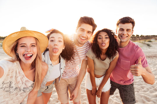 Portrait of happy young multiracial girls and guys 20s in summer clothes taking selfie, while resting at seaside during sunset