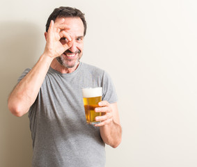 Fototapeta premium Senior man drinking beer with happy face smiling doing ok sign with hand on eye looking through fingers