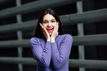 Portrait of  young brunette woman on the gray background of the city wall. she poses, expresses delight and surprise, his mouth wide open. Human emotion.