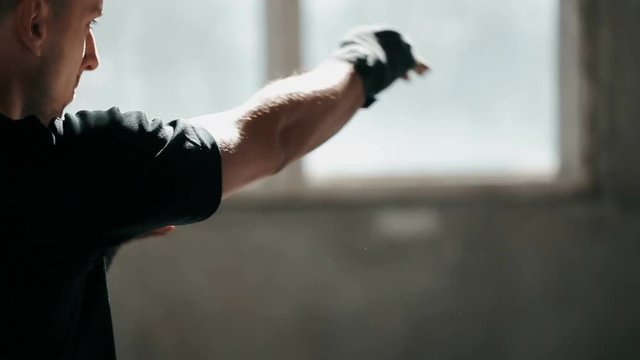 Serious energetic boxer warming up with hitting, wearing black t-shirt and bandage set, keeps fit as doing regular morning exercise