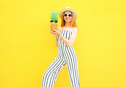 Fashion smiling woman with a pineapple is wearing a white striped pants, round hat on yellow wall background
