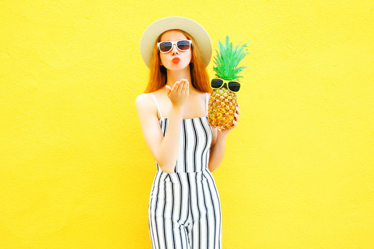Fashion woman sends an air kiss with a pineapple in a white striped pants on yellow background