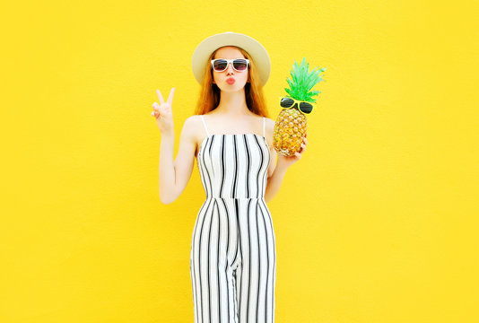 Fashion young woman with a pineapple in a white striped pants, round hat on yellow wall background