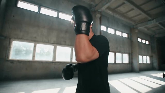 Persistent strong boxer practises different punching techniques, wearing black t-shirt and boxing gloves, having regular training during the week