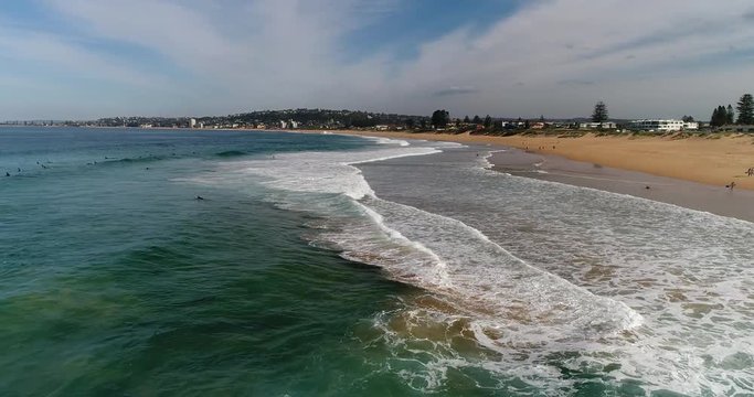 Wide remote sandy Narrabeen Collaroy beach with surfers and rock pool with swimmers in aerial hovering end to end.
