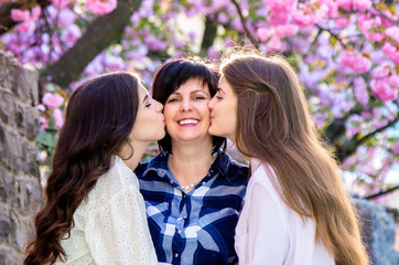 Mother with an adult daughters together kissing elderly mother, in a blooming sakura park