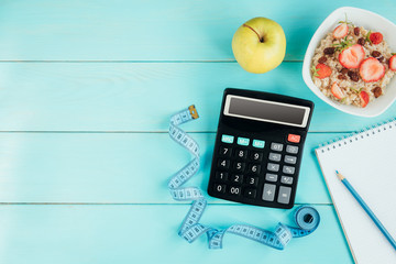 Sneakers, tape measure, notebook, calculator, bottle of water, apple and oatmeal with strawberry and raisins on blue wooden background. Healthy lifestyle, diet concept. Counting calories, menu. - obrazy, fototapety, plakaty
