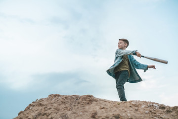 Obraz premium low angle view of boy in denim jacket playing with baseball bat while standing on hill against cloudy sky