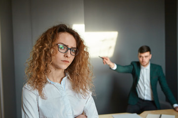 Upset beautiful young woman employee with curly hair and glasses feeling stressed while being fired by her angry boss who in standing in background and pointing finger. Failure, dismissal and firing