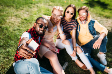 Fototapeta premium selective focus of multiethnic friends taking selfie on smartphone while resting on green lawn in park