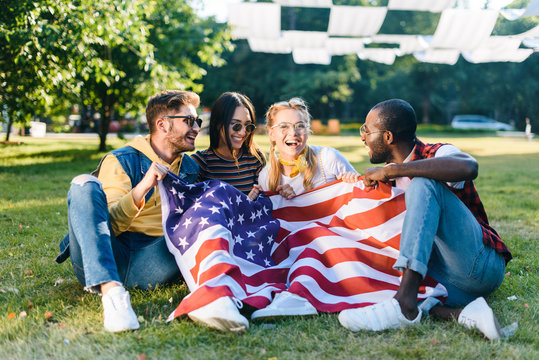 multiracial happy friends with american flag sitting on green grass in park